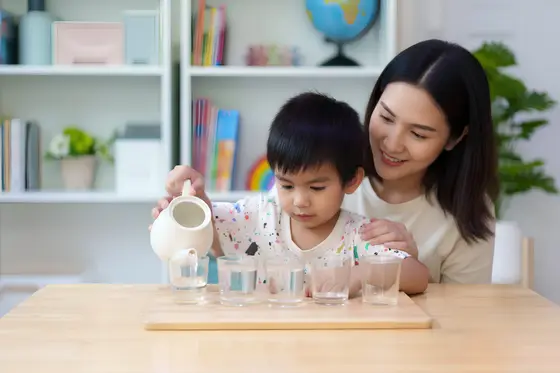 Un enfant verse de l’eau dans des verres avec l’aide de sa mère, activité Montessori de transvasement